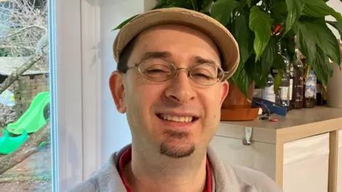 Maurice Chiodo is in the middle of the image smiling. He is wearing a beige cap, glasses and a grey jumper. Behind him is a plant pot on a shelf. 