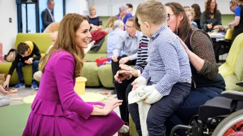 Princess of Wales wears a magenta skirt suit and is kneeling while chatting to a child and his carer. Other adults and children are in the room.