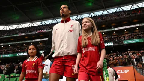 Getty Images Hannah walking next to Virgil van Dijk, in a stadium with fans in the stands behind them. Another girl is on the other side of them. All three are wearing Liverpool kits