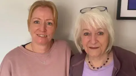 Myton Hospice Two women side by side. The one on the left is blonde, younger and has a pink jumper and necklace with interlinked heart pendant. Her mum is on the right, with bobbed white blonde hair, a purple t-shirt, burgundy fleece and necklace and glasses on top of her head.