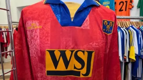 12th Man Retro The image shows a red shirt with the letters WSJ on the front in an orange box. Other football shirts can be seen hanging on a rail in the bacxkground. 