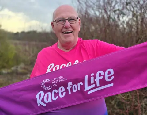 Cancer Research UK A man wearing glasses and a pink top is looking at the camera. He is outside, holding a purple Race for Life banner and bushes and trees are blurred behind him in the photo.