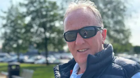 Michael looking at the camera. He is standing outside on a sunny day. He is wearing a navy jacket, blue shirt and sunglasses.