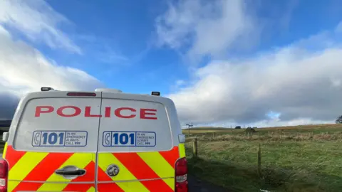 A police van parked next to a field, with a couple of buildings dotted in the distance