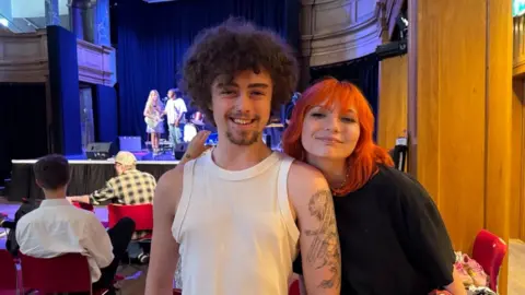Andrew Turner/BBC Dan is wearing a white vest top and has tattoos on his left arm. He has long dark curly hair and a goatee beard. Skar is wearing a black tee shirt and has long orange hair. They are standing in St George's Theatre, where there are people on stage, and an audience on chairs.