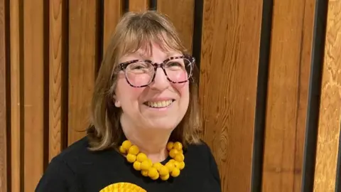 Alison Barnes is smiling at the camera. She has mid-length brown hair and large brown glasses. She is wearing a chunky yellow necklace and a yellow rosette.