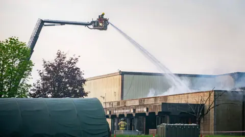 Firefighters using crane at RAF Fairford. They are spraying water onto a building and there is a green tent in the front of the image.
