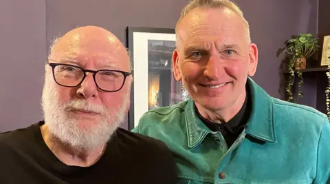 Peter Flannery and Christopher Eccleston standing side by side and smiling into the camera. Flannery is on the left and is bald with a white beard. He is wearing black-framed glasses and a black top. Eccleston is on the right and has grey hair and is wearing a green shirt. They are standing in front of a purple wall which has a photo frame hanging on it. There is a shelf with a plant in the right corner.