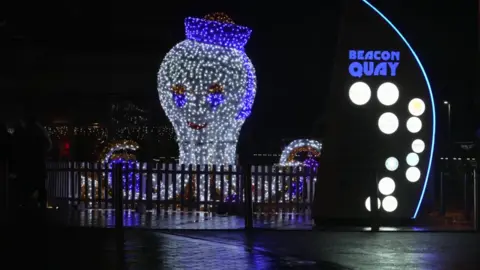 An octopus installation of lights can be seen, with blue light reflected on to the pavement in front of it and a sign reading 'Beacon Quay' to the right.