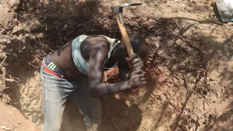 Edwin Naitale / BBC A man crouches in a trench with a hoe as he is about to bring it down to break up the earth 