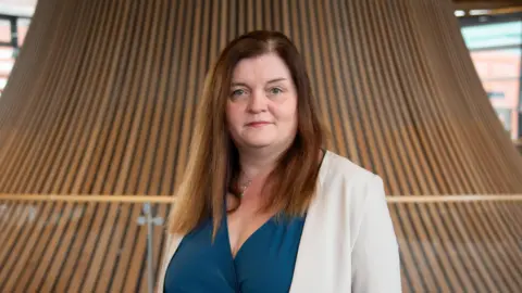 A woman with long brown hair, wearing a blue wrap dress underneath a beige suit jacket stares into the camera in the Oriel in the Senedd.