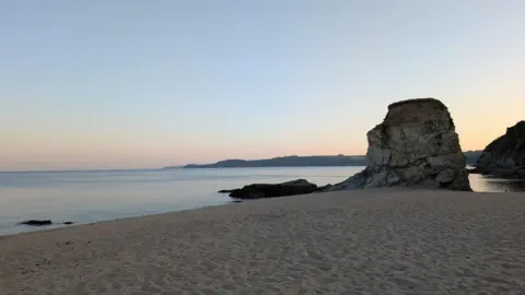 Carlyon Beach at sunset is empty of people but the sand shows many footprints. The sky is pale shades of pink, orange, yellow and blue. There is a large boulder on the shoreline and beyond the sea a headland is in silhouette.
