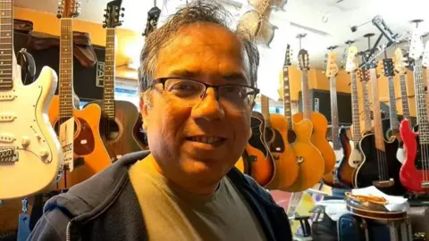 Customer Rajeev Bhattacharyya, visiting the Music Box after having his guitar repaired there. He is looking at the camera, smiling. There are many guitars hanging form the ceiling behind him.