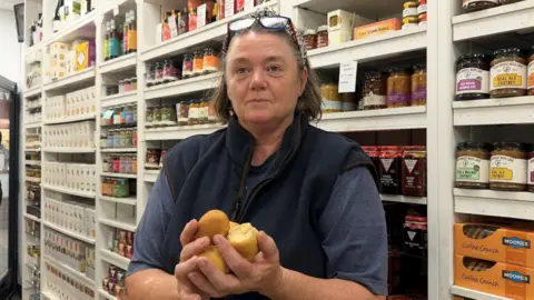 Woman with glasses on her head holding four scone-like round biscuits in both hands. She is wearing a navy body warmer. Behind her are some shelves with some artisan Dorset food products.