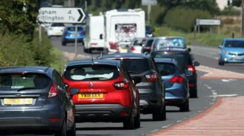 Lots of cars and a couple of campervans are in a long queue of traffic on the A1 in Northumberland. You can also see a sign to the villages of Ulgham and Tritlington