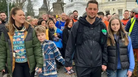 Craig Maxwell with his wife and two kids. There is a crowd of people behind him. His wife is on the let wearing a green puffa jacket with a colourful top underneath and has light brown hair. His son is next to her holding her hand and Craig's hand. He has a checkered top on, Craig is next to him and is wearing a black jacket with black trousers. His daughter is next to him and is wearing a black gillet with a blue jumper underneath. 