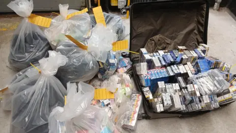 Full plastic bags and a suitcase full of cigarette cartons and vapes on the ground. The bags have been labelled with yellow cards.