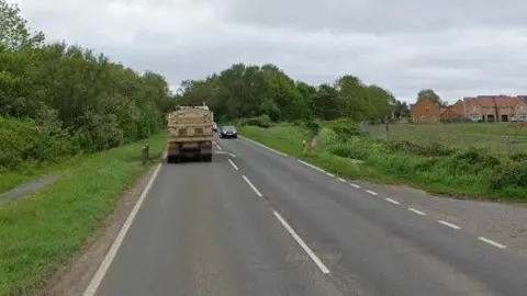 Dual carriageway surrounded by shrubbery and a field. IN the distance are homes while a lorry can be seen on the road.