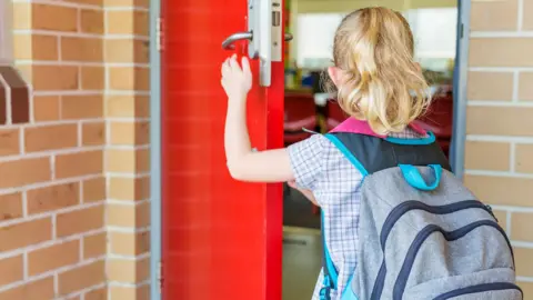 A young girl, with her blonde hair in a ponytail and a rucksack on, opens the door to a classroom