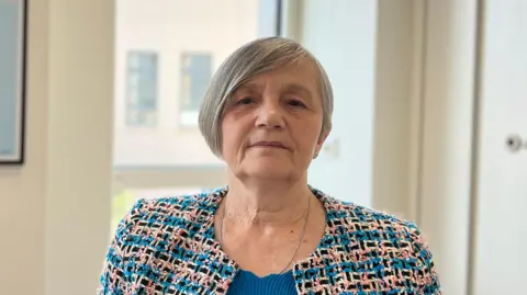 Mary has short grey hair wearing a patterned jacket over a blue top, photographed indoors in an office setting. A framed map artwork hangs on the wall behind, and a window lets in natural light.