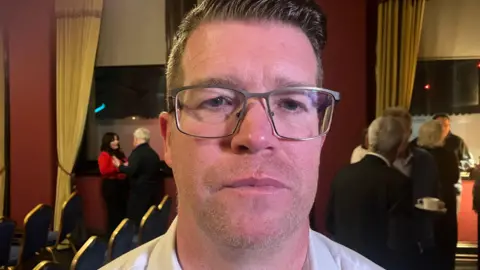 A man with short dark hair wearing grey metal framed glasses