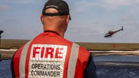 GMFRS A helicopter flies over water in the background as a man wearing a red vest reading 'FIRE OPERATIONS COMMANDER' stands with his back to the camera in the foreground.