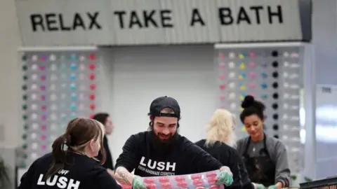 Getty Images A group of men and women making colourful bath bombs in moulds. They are wearing black t-shirts that say Lush. There is a sign on the wall that says Relax Take a Bath.