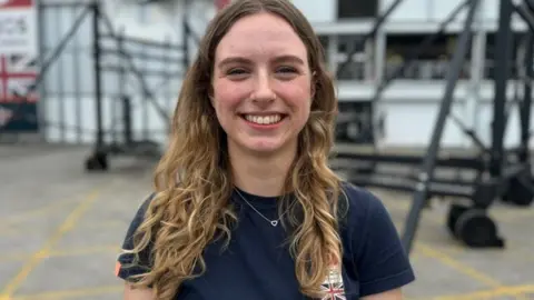 Woman wearing navy blue T-shirt with union jack badge, she has a heart necklace, blonde long wavy hair and is smiling