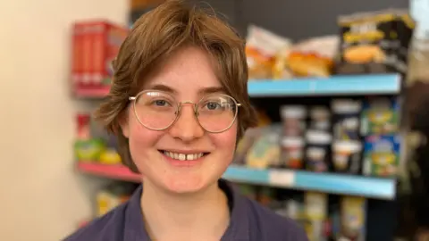 The head and shoulders of Emma Caroe, who is standing in front of shelves of food. Emma has short brunette hair, is wearing rounded silver glasses and is wearing a navy shirt.