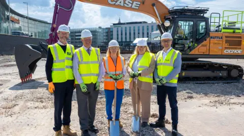 Blackpool Council/MoD Three men and two woman in high- vis vests and hard hats at the ground-breaking ceremony. They stand in front of a large mechanical digger.