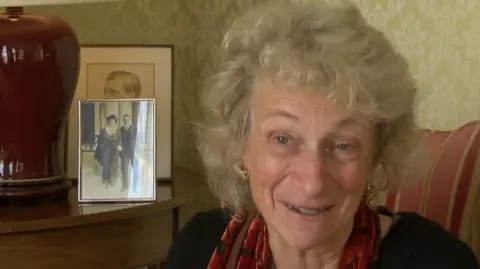 The picture shows a person seated indoors next to a side table with a framed black-and-white photograph of two individuals dressed in formal attire. The table also holds a lamp with a dark red base and a beige shade. Behind the table, there is patterned wallpaper in a muted tone, and part of a striped upholstered chair is visible on the right. The person is wearing a dark top and a patterned scarf in shades of red and black. 
