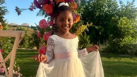 Family handout A smiling young girl is wearing a white dress and stood outside with a flower arch partially seen behind her. 