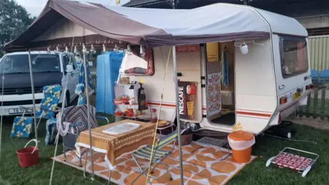The Vanlife Festival A white caravan sits on a patch of grass with a white awning. An orange and white rug has been laid on the grass with table and chairs.