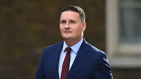 Getty Images A shot of Wes Streeting from the chest up. He wears a navy suit with a light blue shirt and a maroon tie, and his hair is short. Behind him the background is blurred but he appears to be outside and mid-stride. His eyes are looking to the left of the camera.