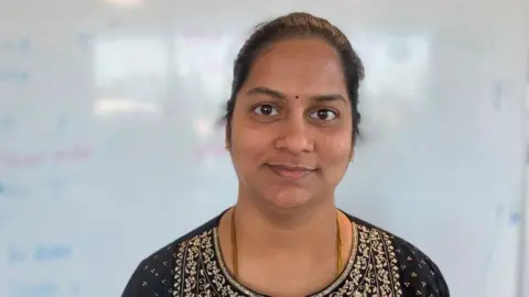 Seetha has her hair slicked back into a bun, a gold thread chain, wearing a black and jewel pattern Salwar Kameez. Wearing gold earrings and a nose piercing and has a dark red bini in between her eyebrows. 