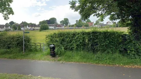 Google A public footpath sign pointing across a grassy field next to a metal gate and a black litter bin next to it. Houses are in the distance. 
