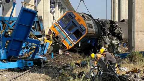 A damaged train lying on its side surrounded by twisted metal with overhead rail tracks in the background