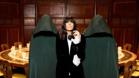 Claudia Winkleman in a black velvet suit flanked by figures dressed in dark green cloaks in front of a large round table