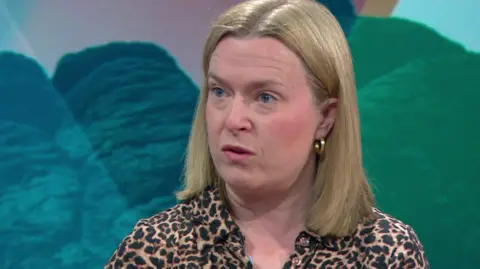 A mid-shot of Rebecca Smith sitting in a TV studio. She has shoulder length brown hair and is wearing a leopard print top.