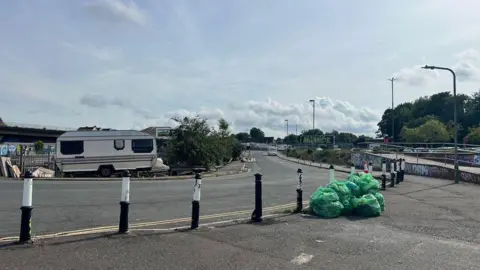A view of New Stadium Road with two parked caravans on it. There is a pile of green rubbish bags on the pavement and some other flytipped materials to the left by one of the caravans.