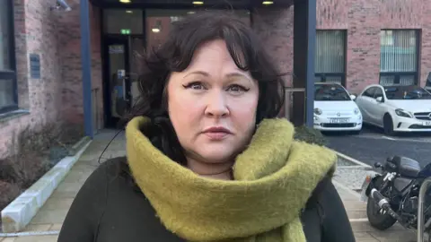 BBC Sara Morrison stands outside the entrance to a red brick municipal building. Her shoulder-length dark hair is parted in the middle and her expression is neutral. She is wearing a black long-sleeved top and her green woollen scarf is around her neck and over her shoulder. 