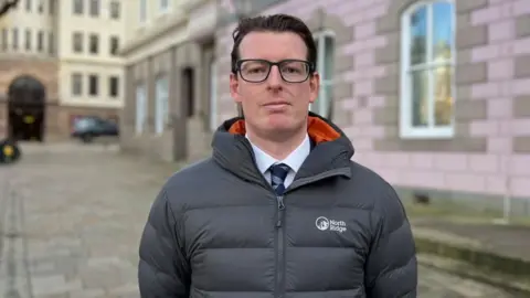 A man with slicked black hair and black-rimmed glasses looks into the camera. He is wearing a grey coat with an orange hood lining over a a suit with a white shirt and navy and light blue striped tie.