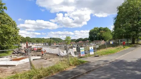 A piece of land separated from the road by metal fences. There are construction signs on the fences. Inside is various construction equipment, including an orange digger, two builders in orange jackets, and white tarpaulin.