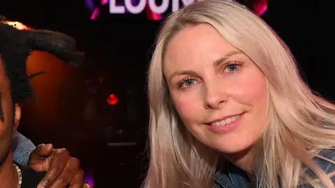 Charlie Hedges with long blonde hair smiling at the camera against a dark background. The bottom of the word "Lounge" in white letters is visible behind her.