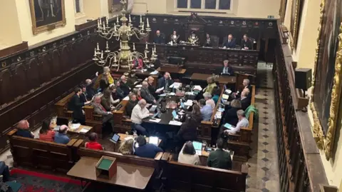 Exeter City Council members sitting in the Guildhall in Exeter with the Mayor of Exeter seated in front of a mace overseeing the meeting.