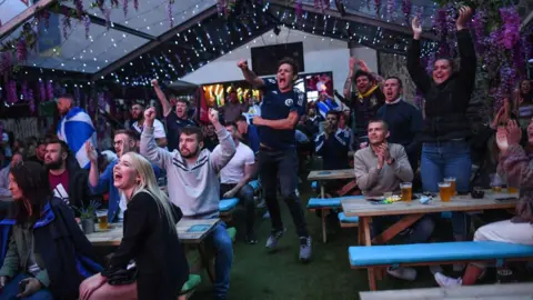 Football fans watching a game while in a pub's beer garden, with drinks  on tables in front of them.