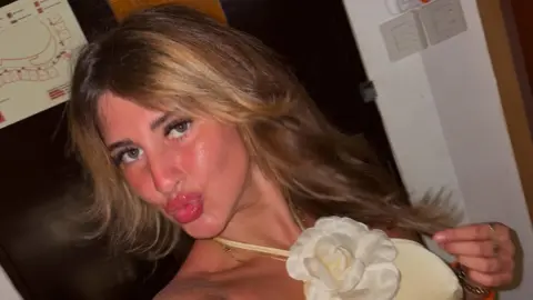 Bella Culley A selfie taken by Bella Culley of her wearing a yellow dress with a fabric white flower at her neck She is pouting at the camera and holding a strand of hair in her other hand.