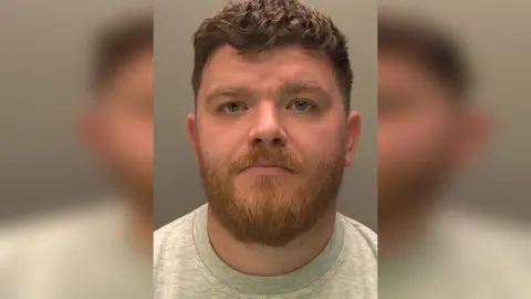 Lincolnshire Police Police custody photograph of Stuart Wells who has dark short hair and beard and is wearing a grey shirt