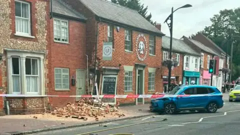Archie Voyde A damaged blue car is parked near a partly damaged red-brick building. A pile of fallen bricks and debris covers part of the pavement.