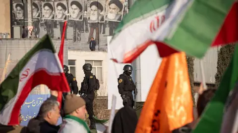 Getty Images Police officers in all black clothing carrying guns patrol a crowd carrying Iranian flags.